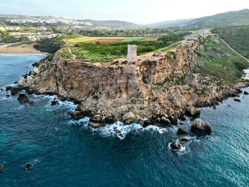 Vue aérienne de la Tour de Għajn Tuffieħa perchée sur une falaise rocheuse surplombant la mer Méditerranée, avec des terres agricoles verdoyantes en arrière-plan à Malte