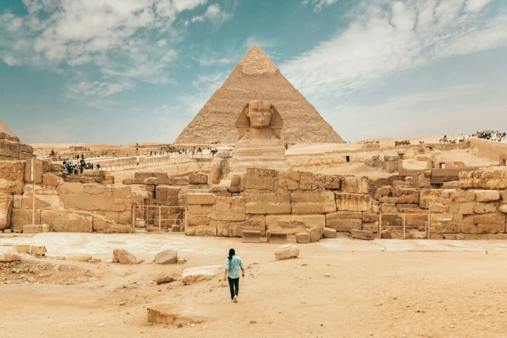 Une voyageuse marchant vers le Sphinx de Gizeh avec la pyramide de Khéphren en arrière-plan, sous un ciel partiellement nuageux