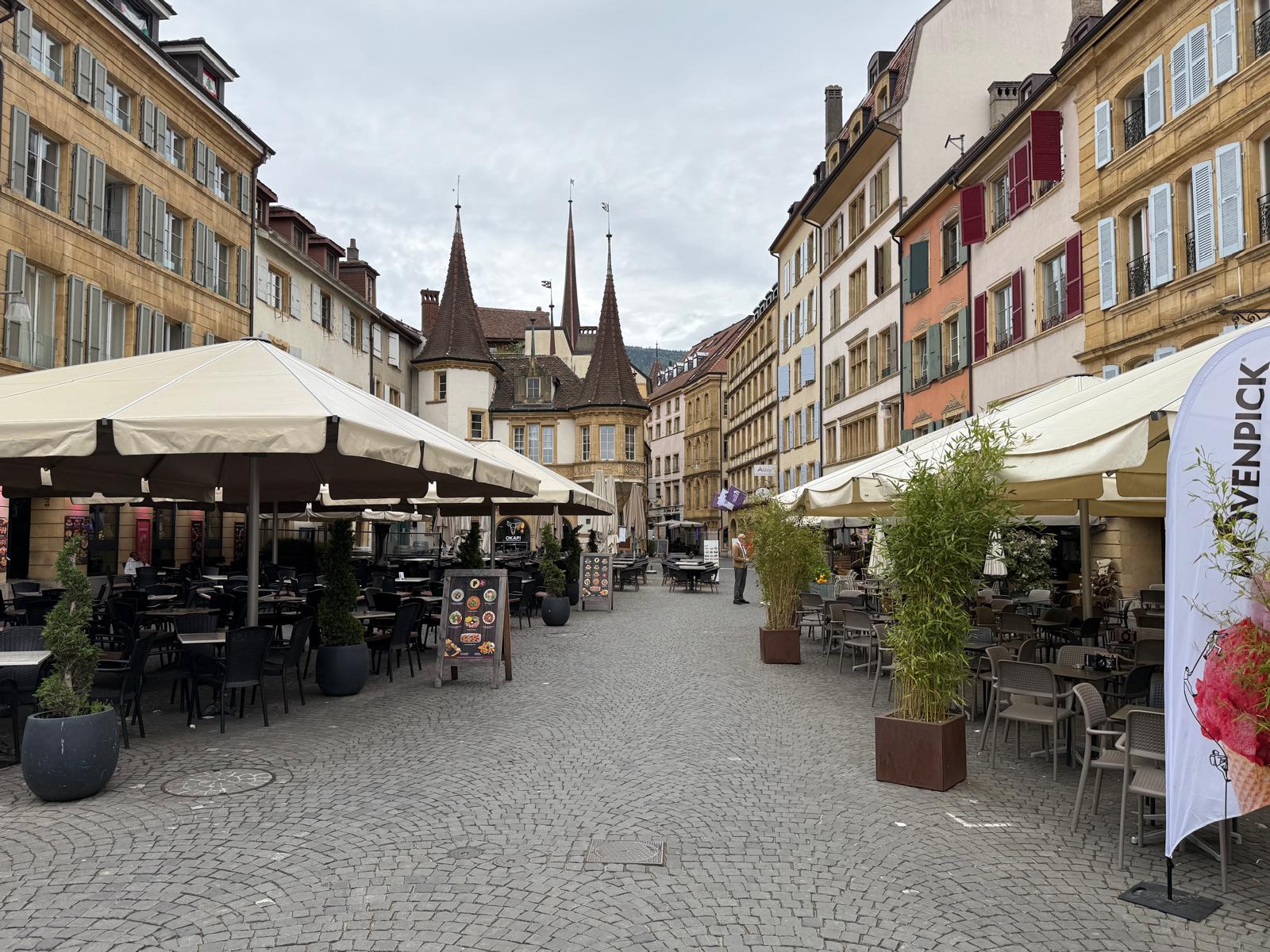 Vue sur les pavés et les grandes terrasses de cafés de la Place des Halles à Neuchâtel, avec la célèbre Maison des Halles et ses tourelles historiques en arrière-plan sous un ciel nuageux.