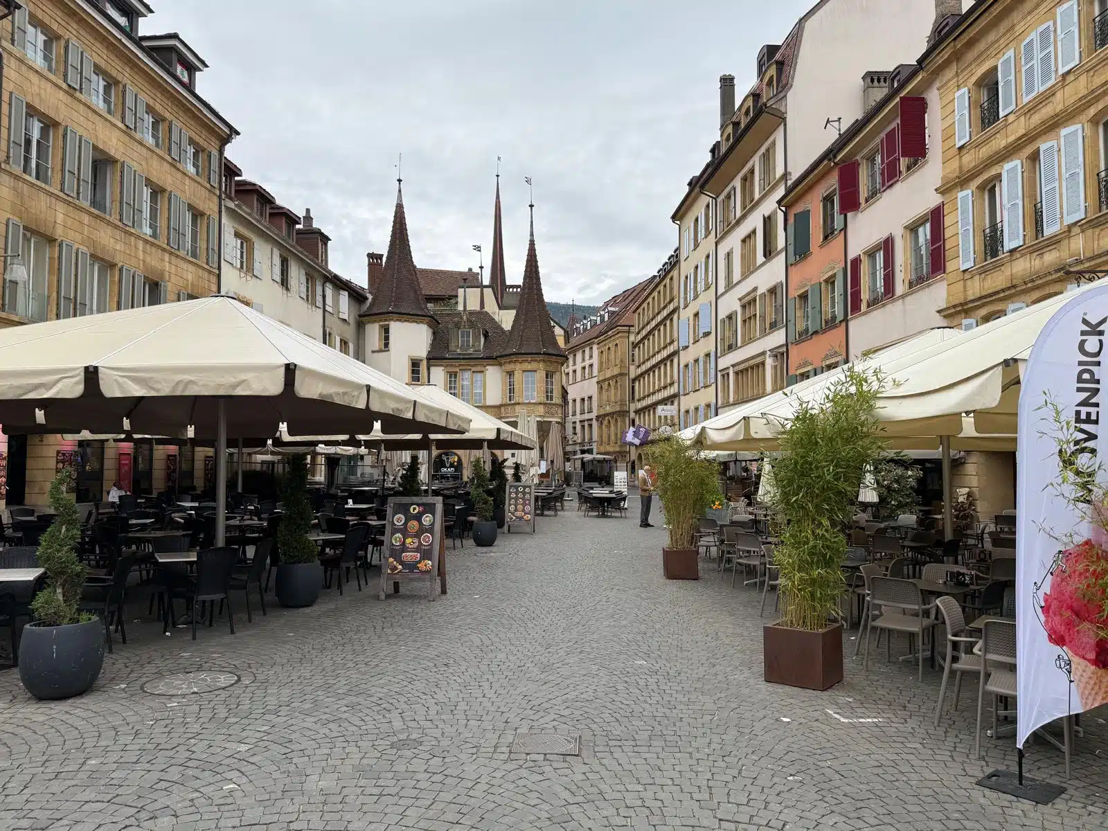 Vue sur les pavés et les grandes terrasses de cafés de la Place des Halles à Neuchâtel, avec la célèbre Maison des Halles et ses tourelles historiques en arrière-plan sous un ciel nuageux.