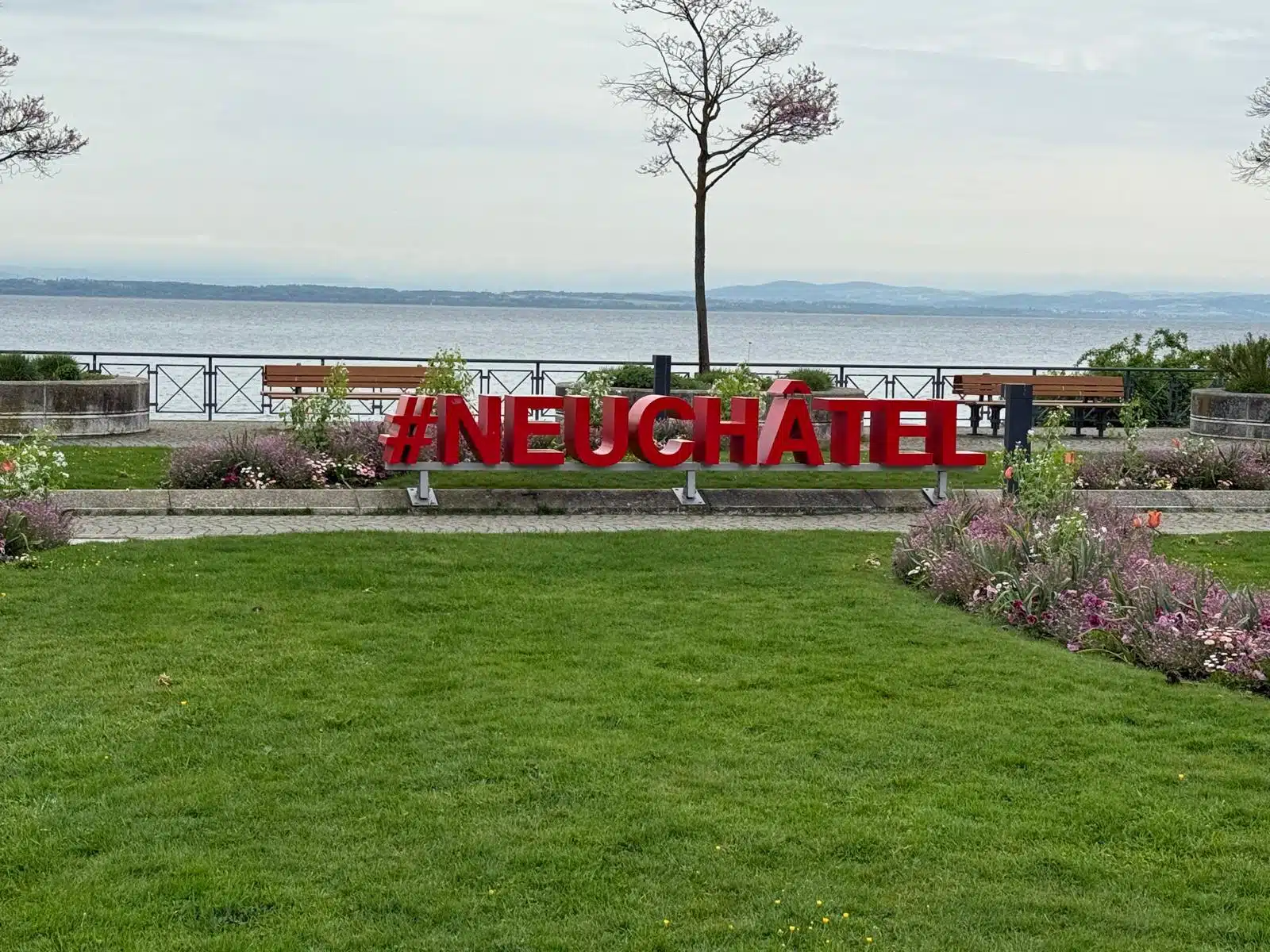 Grandes lettres rouges en 3D formant le hashtag #NEUCHÂTEL posées sur une pelouse verdoyante, avec les eaux calmes du lac de Neuchâtel et les montagnes en toile de fond.