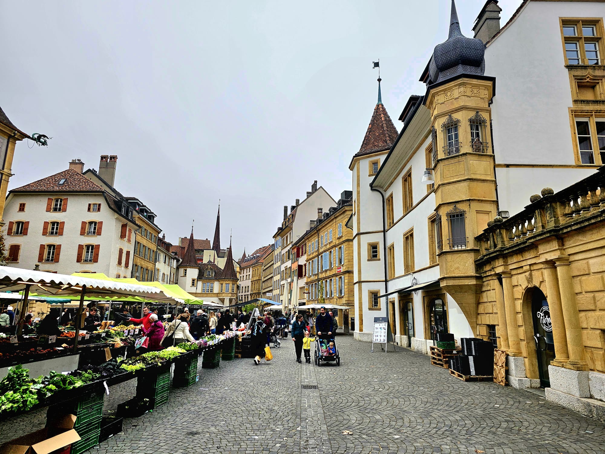Ambiance animée du marché traditionnel sur la Place des Halles à Neuchâtel, avec des étals de produits frais alignés sur les pavés, entourés d'édifices historiques à tourelles sous un ciel gris.