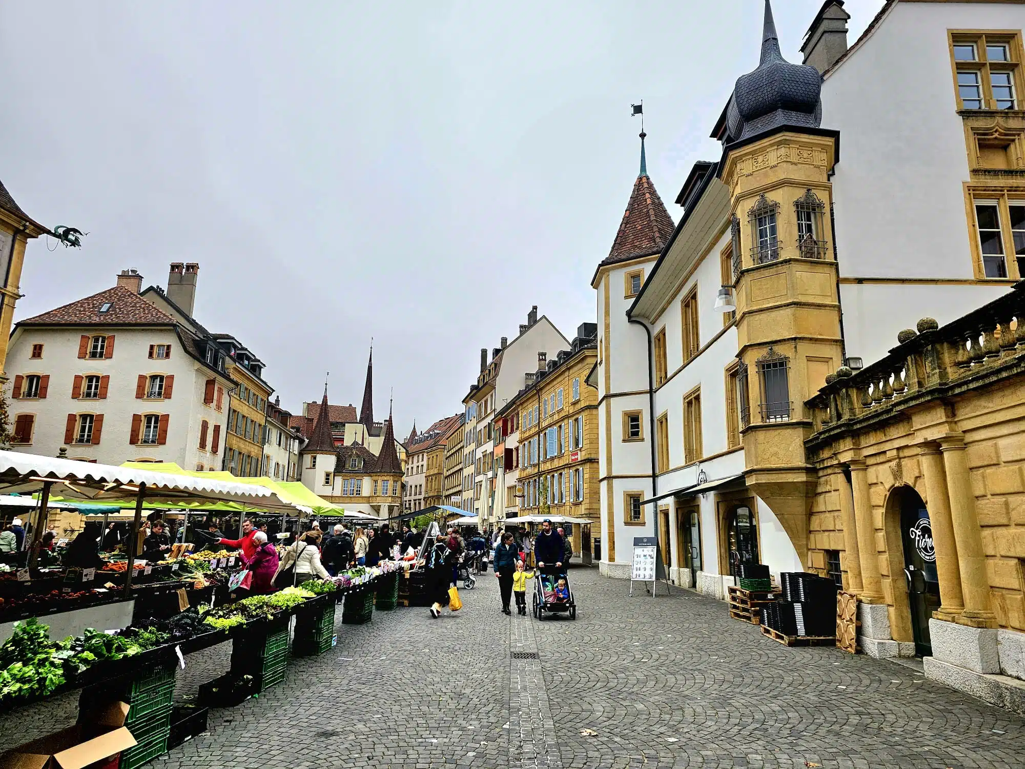 Ambiance animée du marché traditionnel sur la Place des Halles à Neuchâtel, avec des étals de produits frais alignés sur les pavés, entourés d'édifices historiques à tourelles sous un ciel gris.
