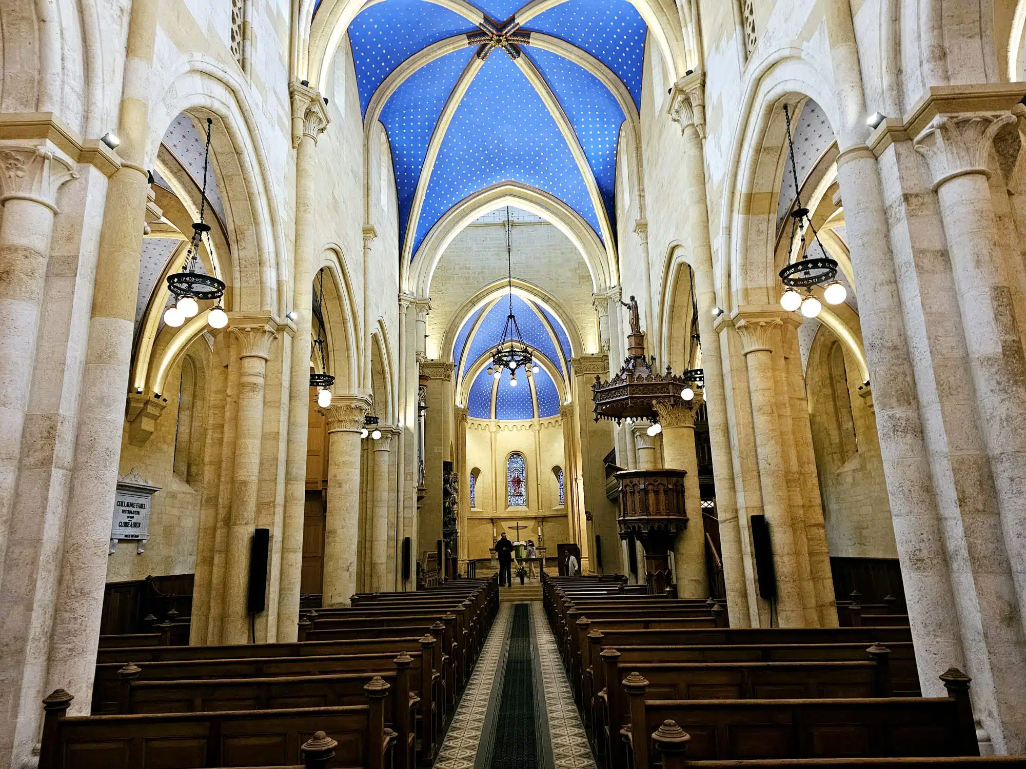 Vue intérieure de la nef de la Collégiale de Neuchâtel, révélant ses piliers en pierre claire, son mobilier en bois sculpté et son impressionnant plafond voûté bleu azur constellé d'étoiles.