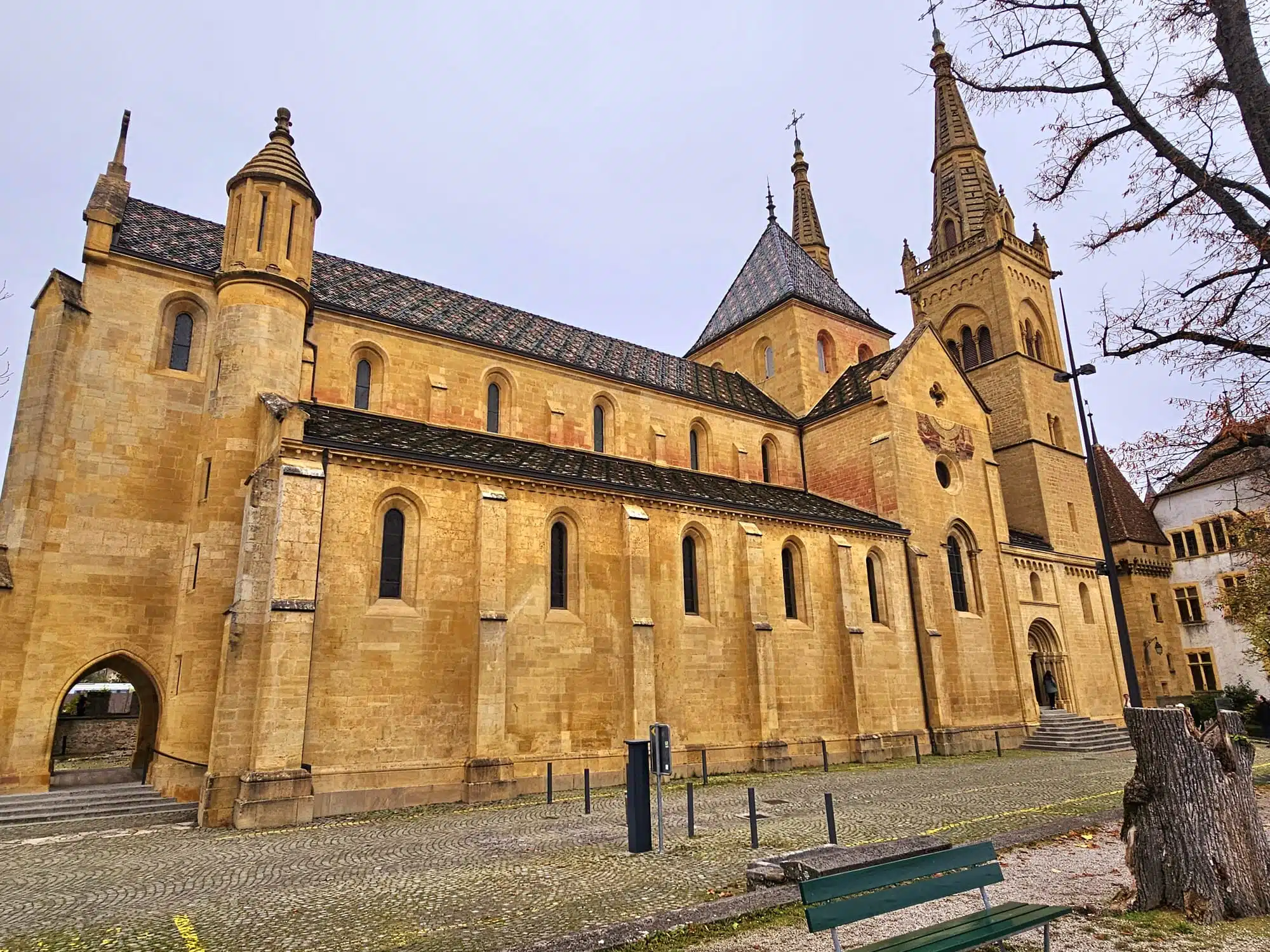 Vue extérieure de la majestueuse Collégiale de Neuchâtel, mettant en valeur son architecture en pierre jaune d'Hauterive et sa toiture aux tuiles vernissées colorées.