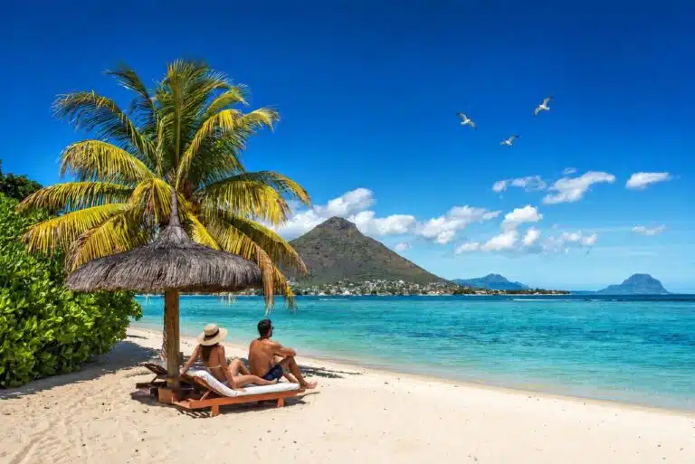 Couple de touristes se relaxant sur une plage de l’île Maurice face à un lagon turquoise et aux montagnes, avec trois oiseaux marins dans le ciel.
